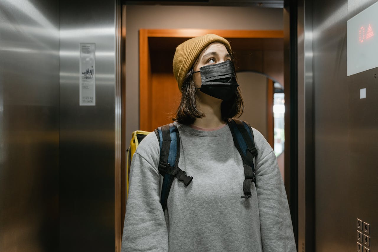A delivery person with a backpack waits in an elevator, wearing a mask and beanie.